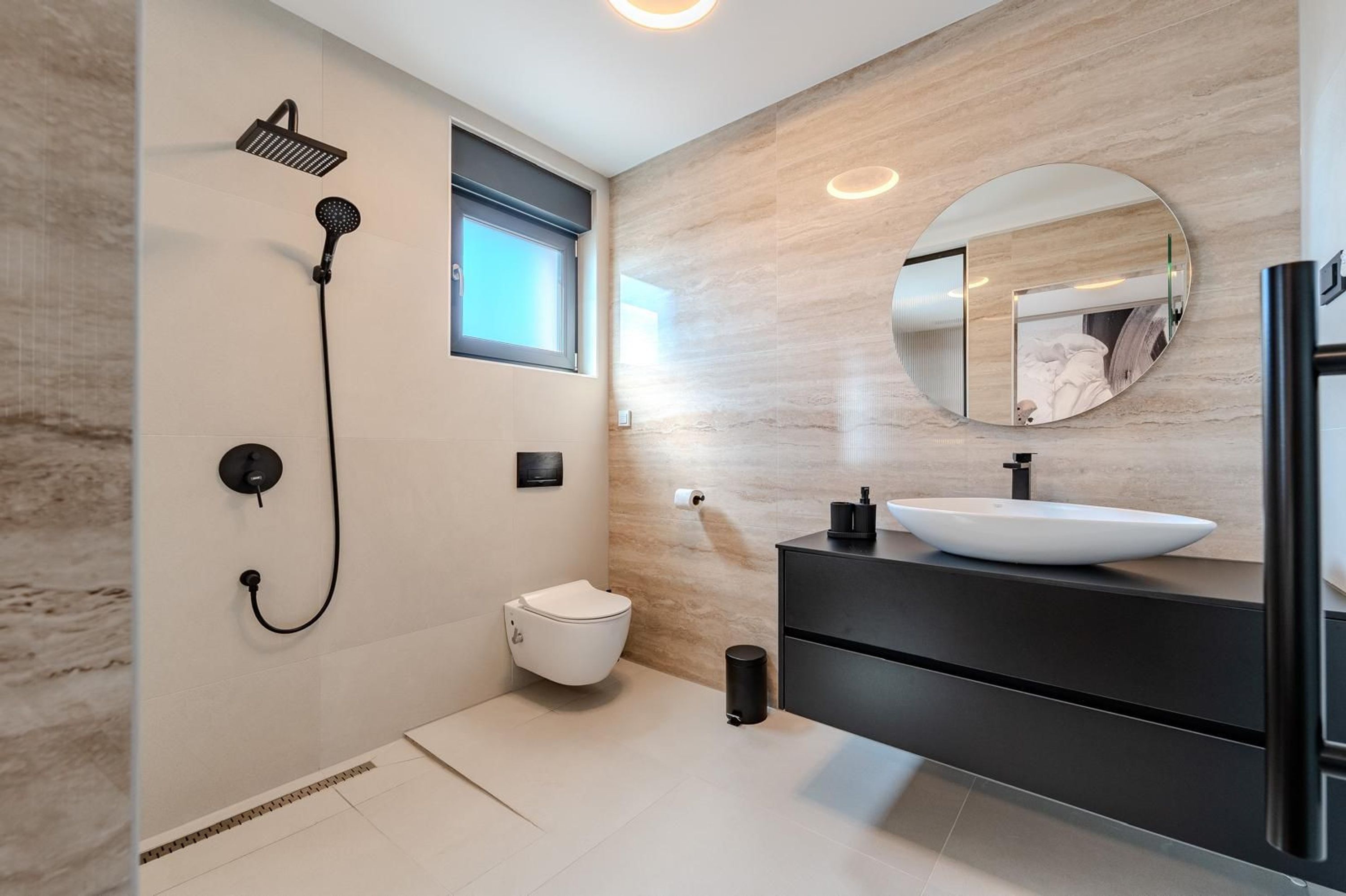 Et moderne badeværelse med toilet i hvid, sort vask, rundt spejl og brusearmatur.