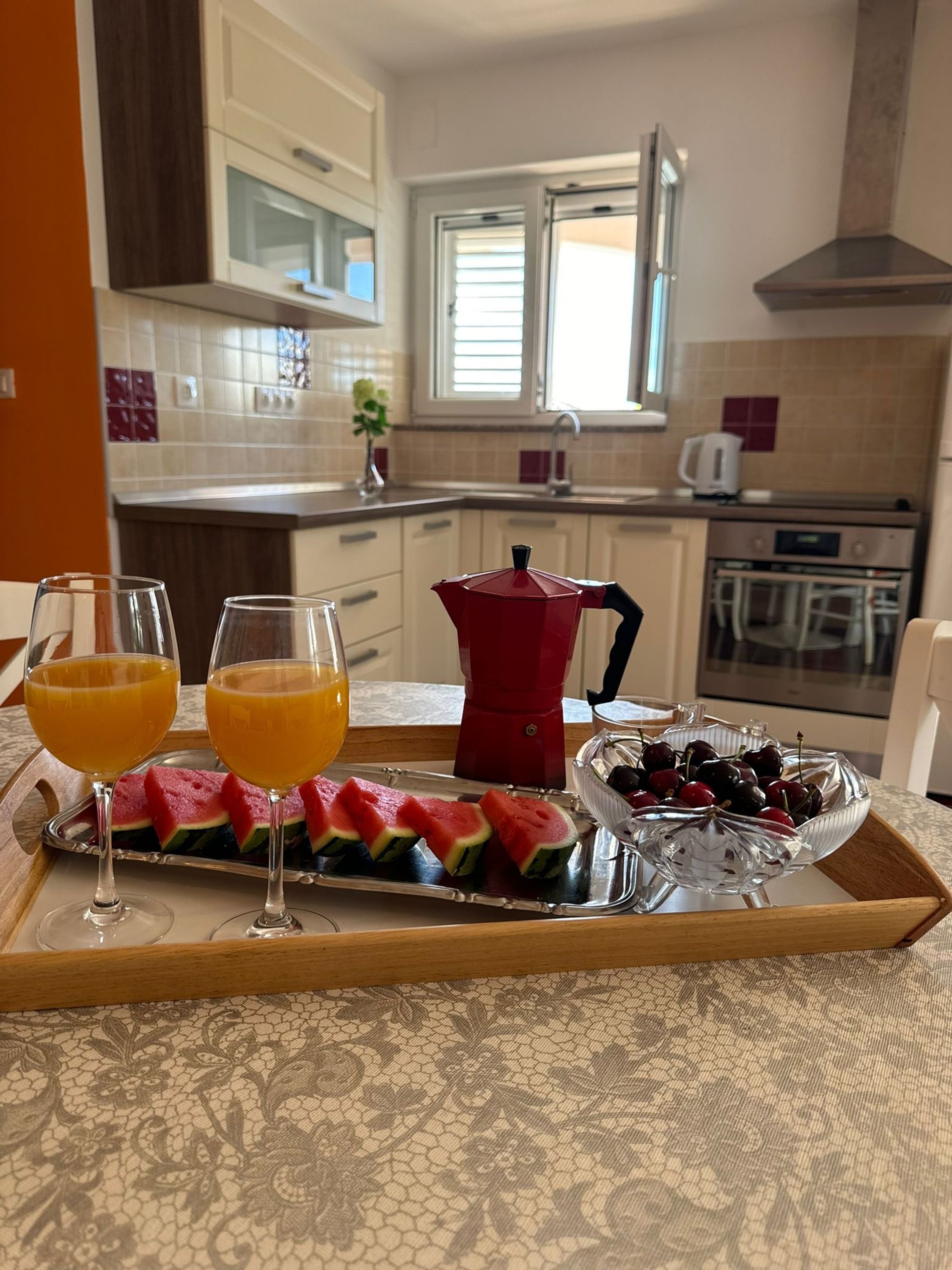 A breakfast tray with watermelon slices, cherries, and orange juice glasses on a table in a kitchen with white cabinets and a window.