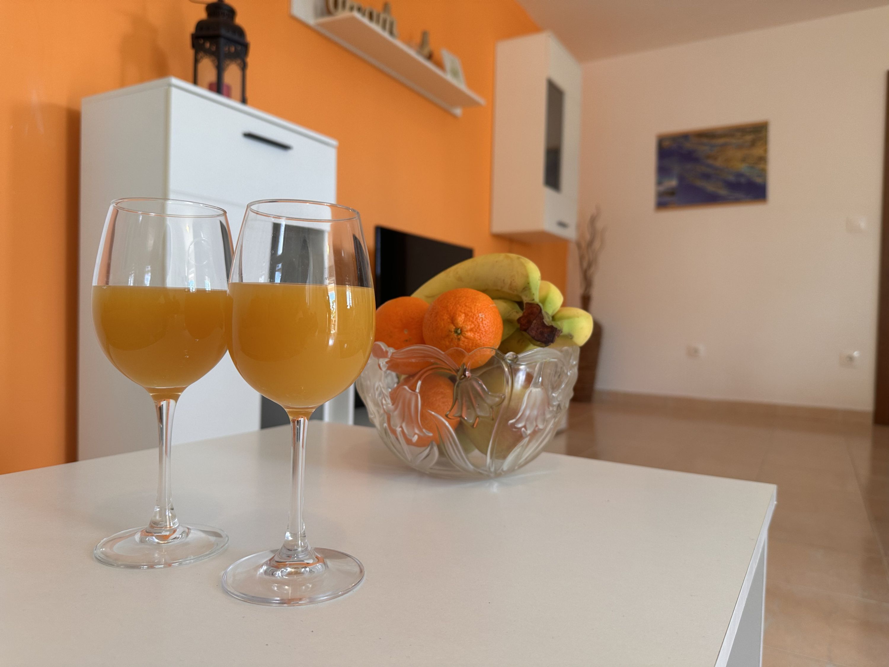 Two glasses of orange juice on a table with a fruit bowl containing oranges and bananas.