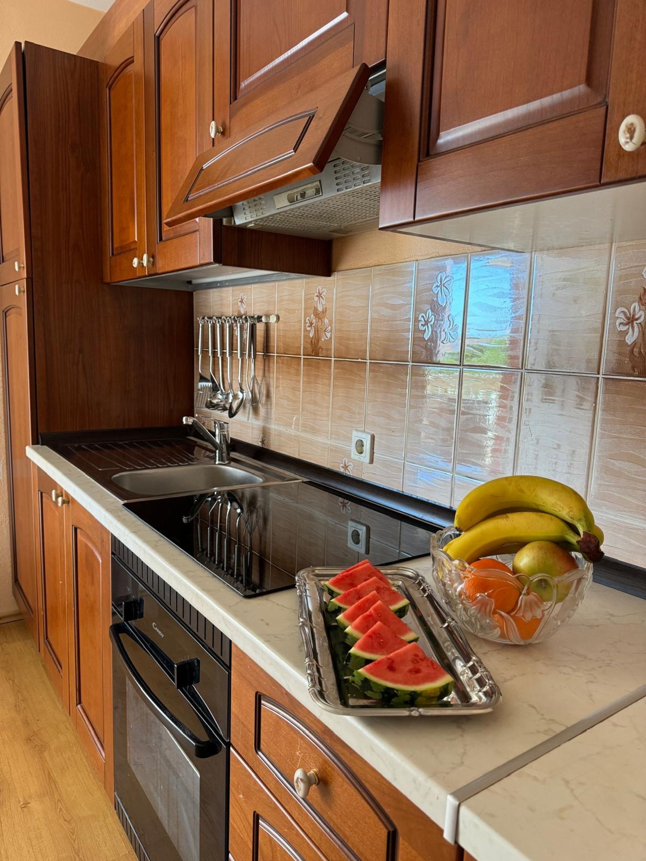 Eine Küche mit Holzschränken, einer Edelstahl-Spüle und einer Arbeitsplatte mit einer Obstschale, die Bananen, Äpfel und Wassermelonenstücke enthält.