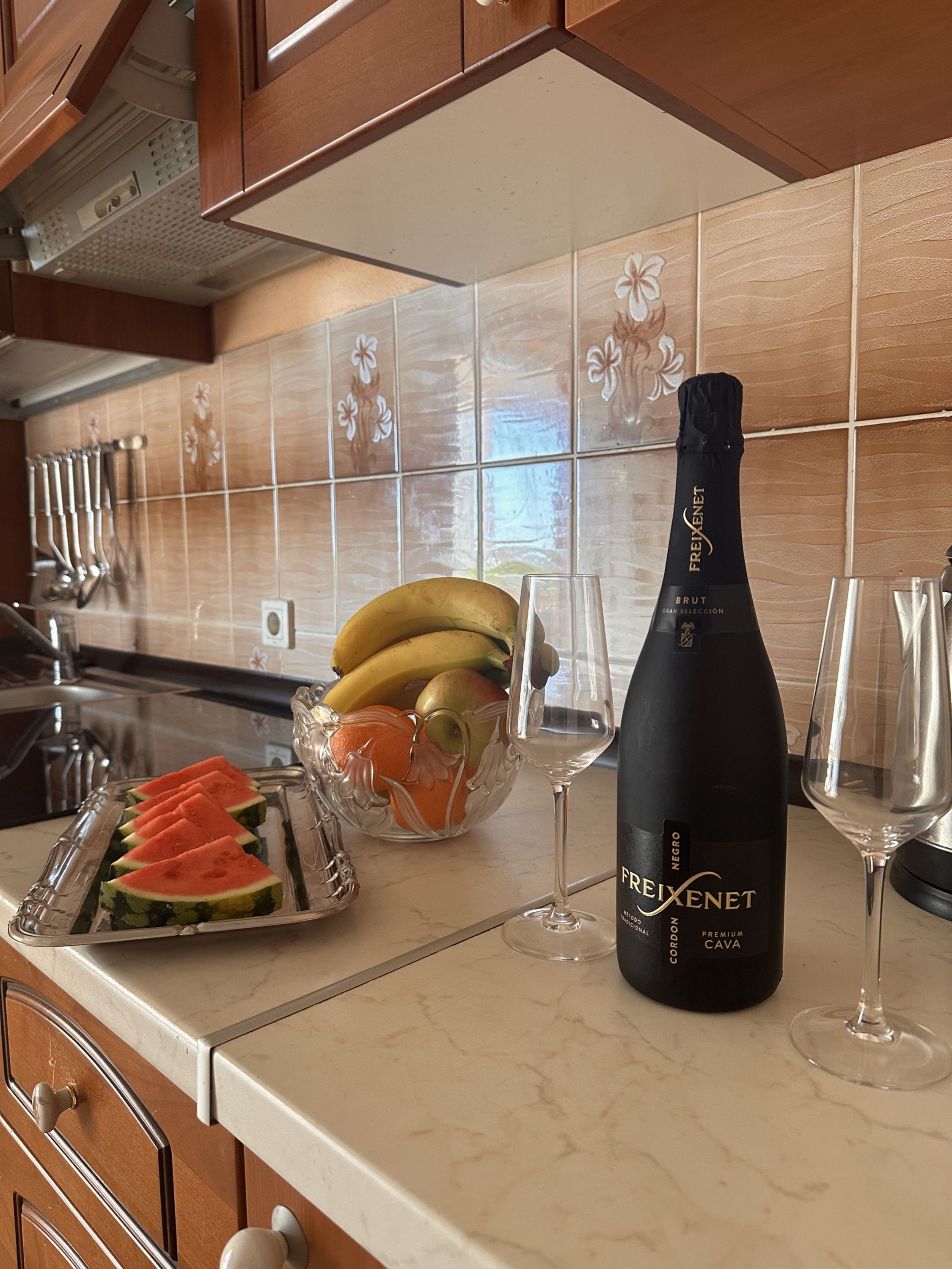 A kitchen counter with a bottle of Freixenet Brut Cava, a glass of wine, and a fruit bowl containing bananas and oranges.