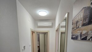 Een smalle gang met een plafondlamp, een airconditioner en een deur naar een andere kamer.