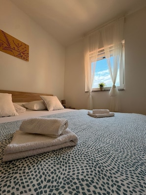 A hotel room with a bed, pillows, a nightstand, a window with sheer curtains, and a small potted plant. - Beautiful Apartment Bodulo 6 (Povljana)