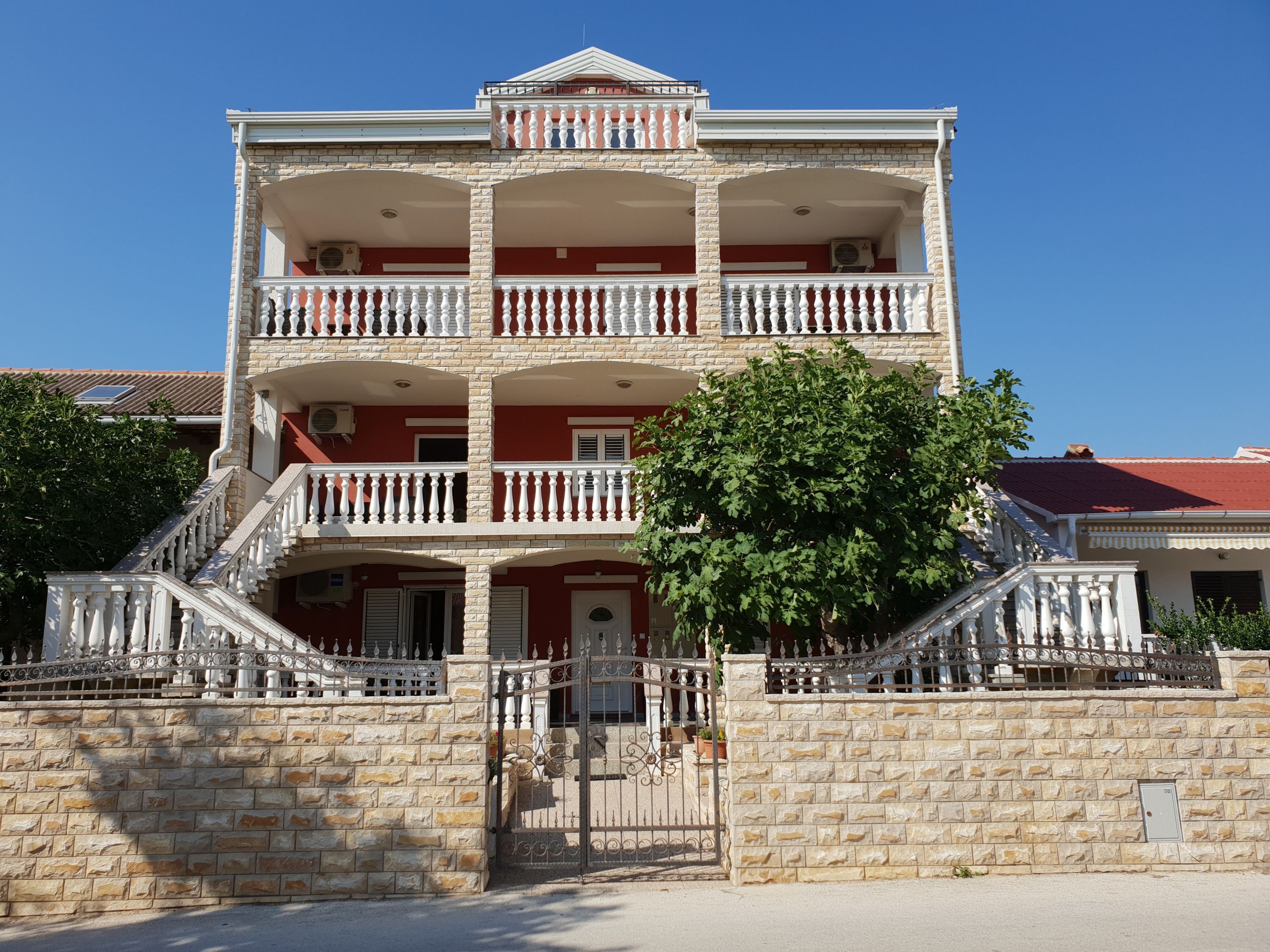 Un edificio de varios pisos con una escalera central, balcones y un muro de piedra que rodea la propiedad.