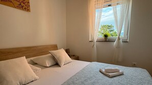 Una cama bien hecha con una colcha estampada, dos almohadas y una toalla doblada. Se ve una ventana con cortinas transparentes y vistas al exterior con vegetación.