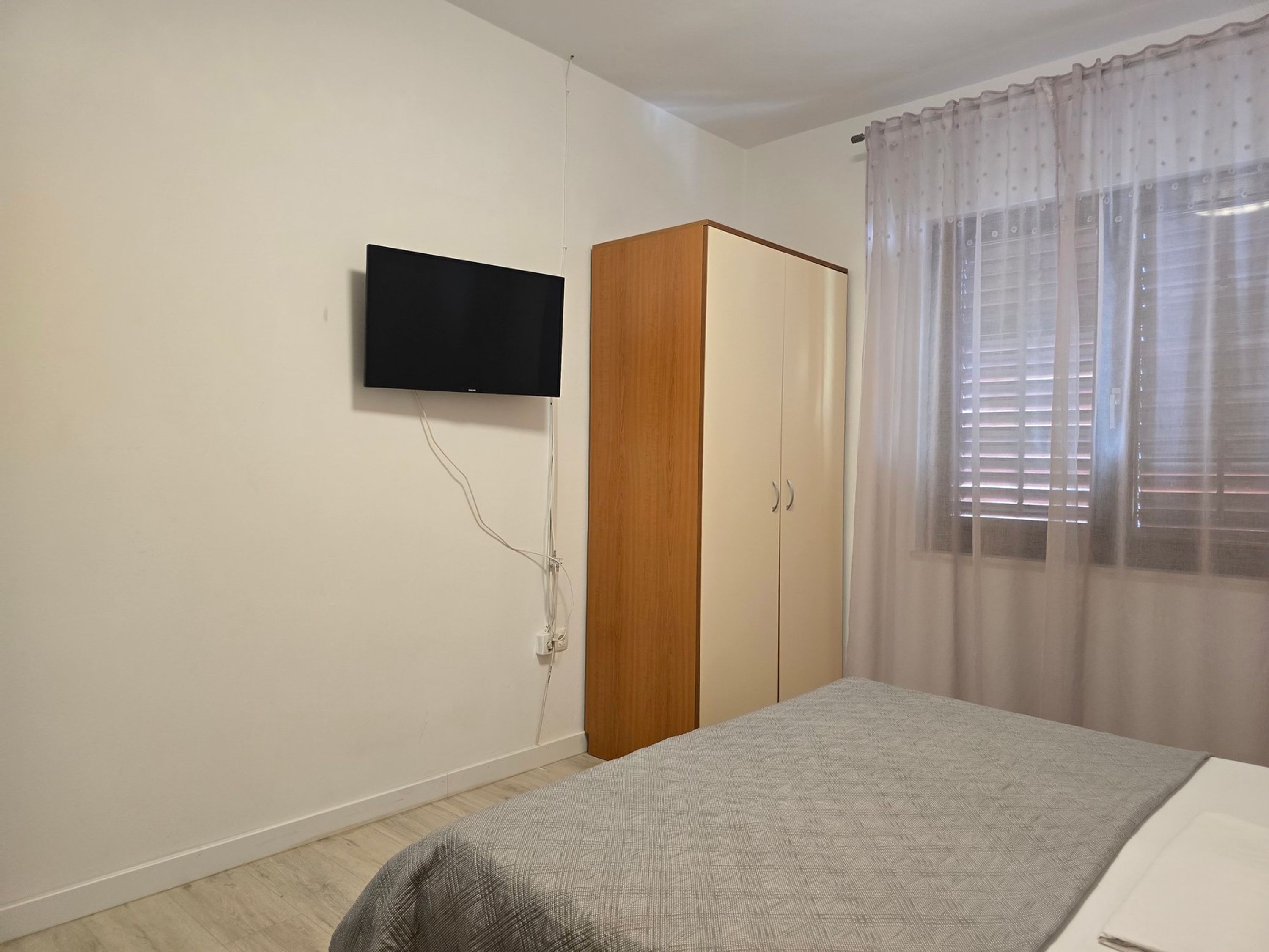 Een slaapkamer met een bed, een kledingkast, een aan de muur bevestigde televisie en een raam met gordijnen.
