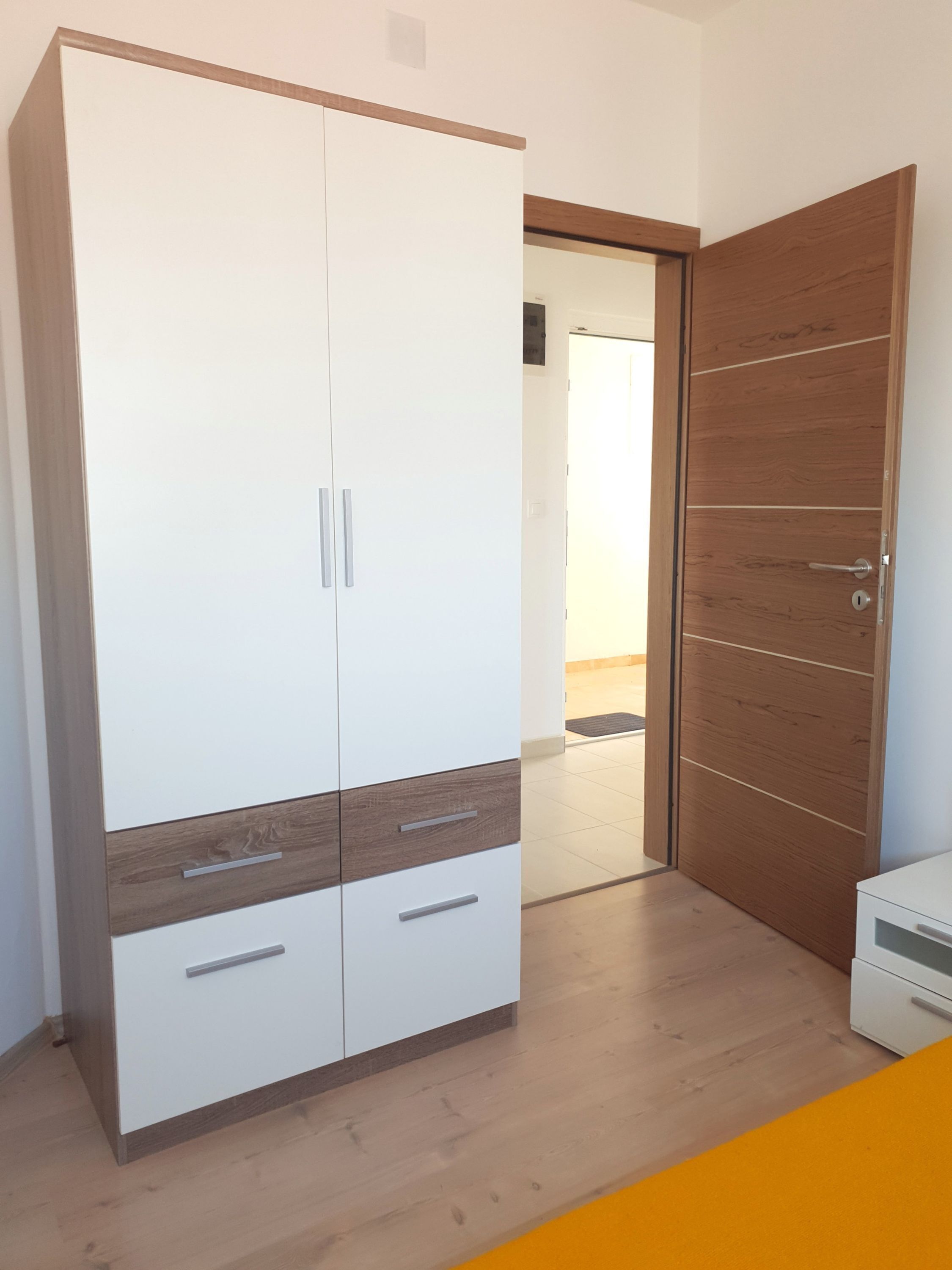 A room with a wardrobe featuring two doors and a wooden panel design, a door with a handle, and a small white cabinet.
