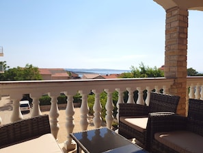 A balcony with wicker furniture and a glass table, overlooking a street and distant buildings. - Beautiful Apartment Bodulo 5 (Povljana)