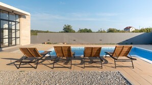 Un espace piscine extérieur moderne, doté de quatre chaises longues en bois et surplombé d’un ciel bleu azur.