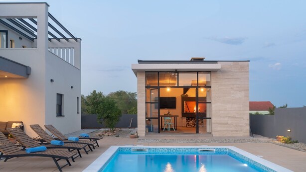 Un espace moderne aménagé au bord de la piscine, avec une piscine rectangulaire, des chaises longues et un bâtiment clos par des parois de verre, doté d’une cheminée.