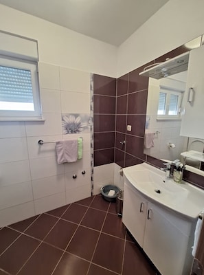 Ein Badezimmer mit weißem Waschbecken, einem Spiegel, einem Handtuchhalter und einem Fenster mit Jalousien.
