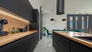 A modern kitchen with dark cabinetry, a wooden countertop, and a built-in oven. There is a dining area with a teal chair and a plant. - Modern Villa Porta in Novalja (Novalja)