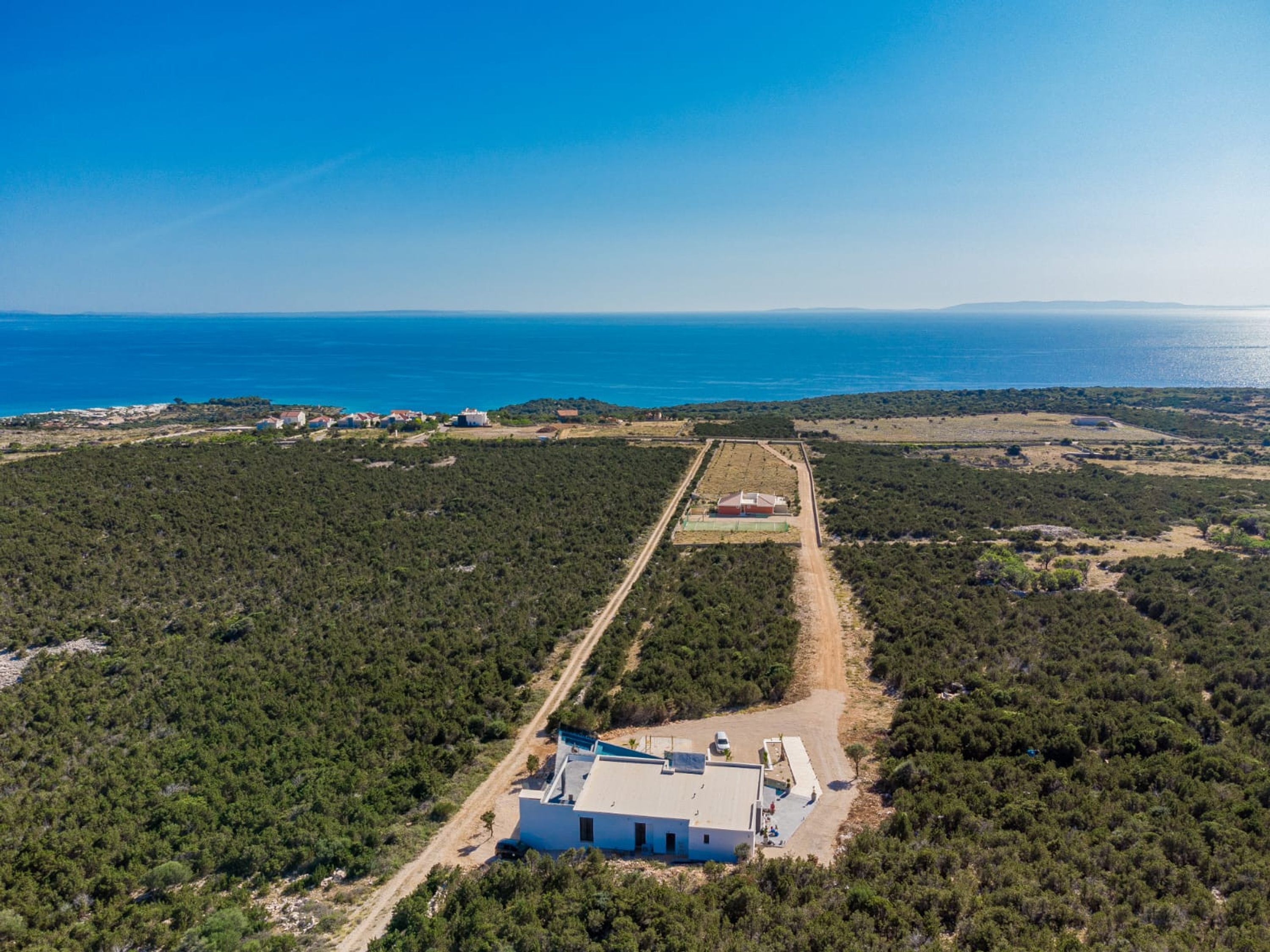 An aerial view of a coastal property with a large building, surrounded by dense forest and a clear view of the sea.