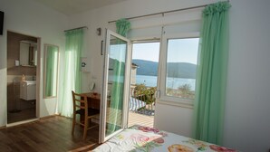 Et soveværelse med trægulv, seng med blomstret sengetæppe, stol, skrivebord, badeværelse med spejl og en balkon med udsigt over vand og bjerge.
