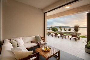 Uma sala de estar moderna com um sofá, uma mesa de centro de madeira com uma bandeja de frutas, e vista para uma piscina e espreguiçadeiras.