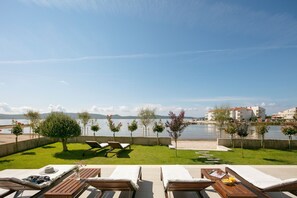 Uma área à beira da piscina com espreguiçadeiras e uma mesa, com vista para um corpo de água e edifícios ao longe.