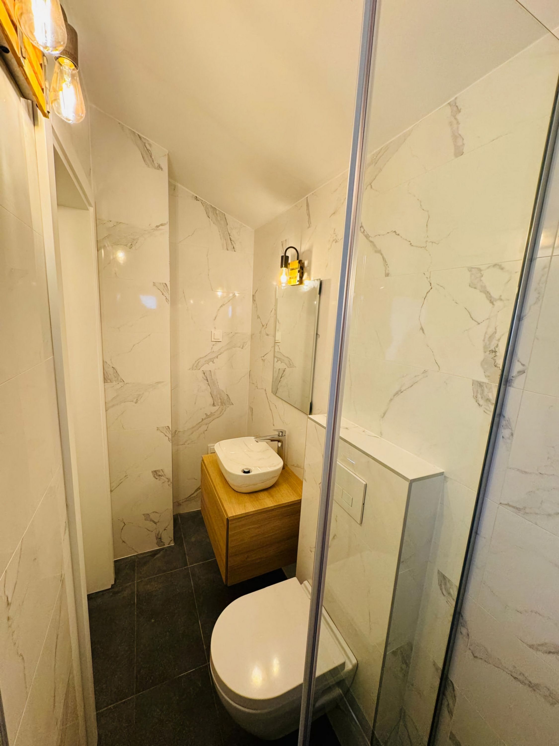 Ein modernes Badezimmer mit Marmorwand, Waschtisch aus Holz, weißem Waschbecken und einer Duschkabine aus Glas.