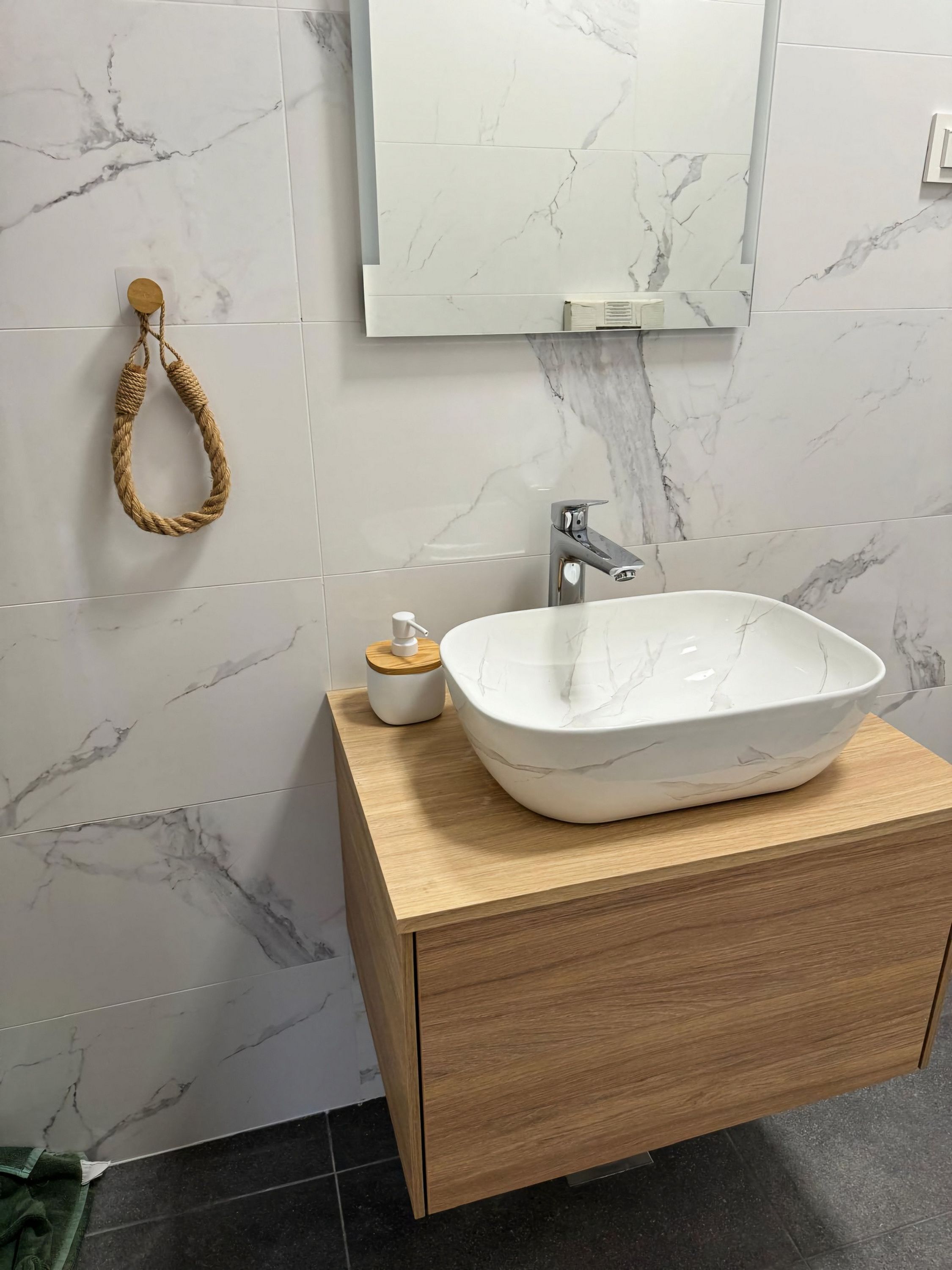 A bathroom with a marble-tiled wall, a wooden vanity, a white marble sink, a towel ring, and a mirror.