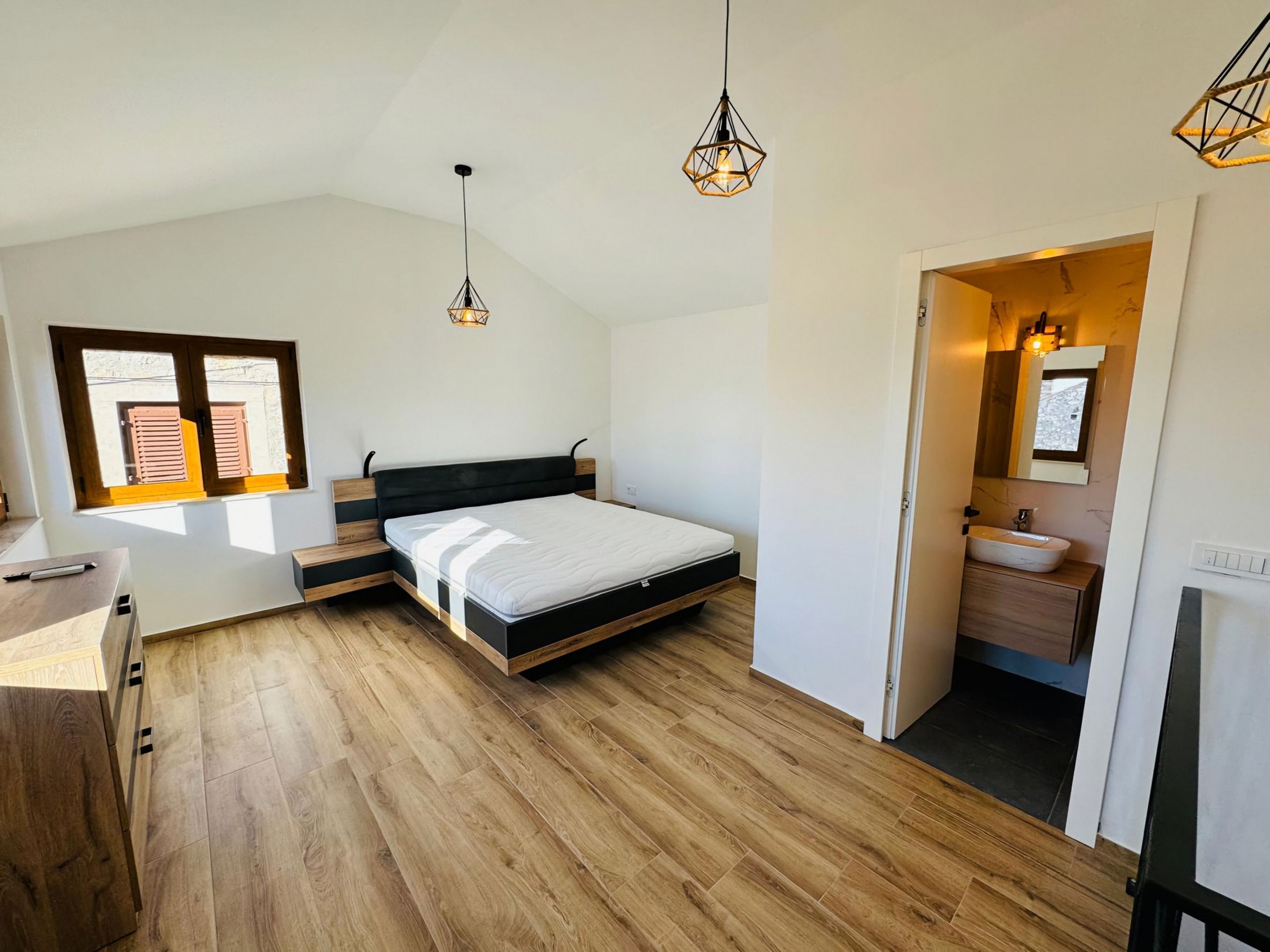 A modern bedroom with a wooden floor, a bed with a black headboard, a desk, and a bathroom with a sink and mirror.