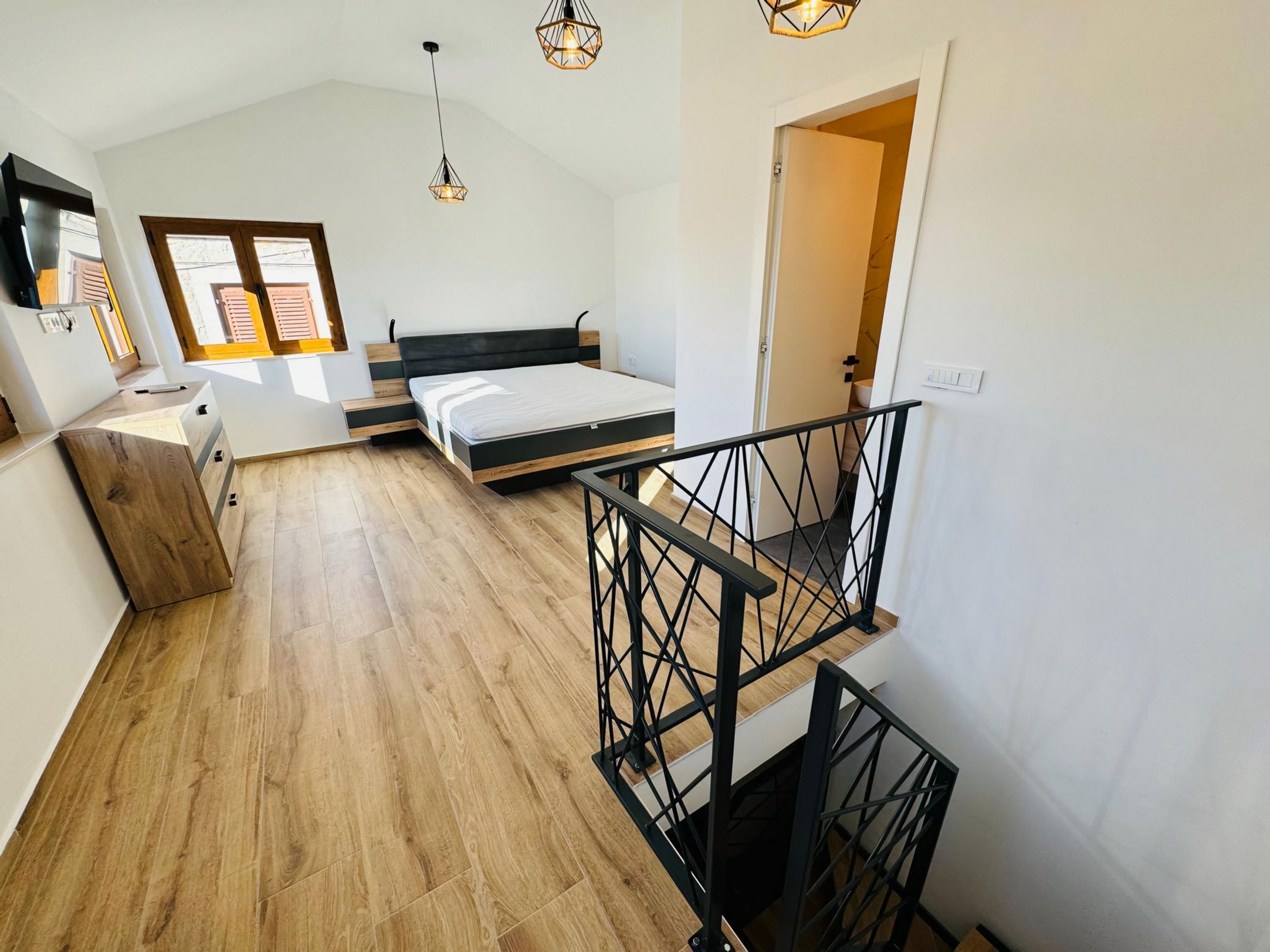 A modern loft bedroom with a wooden floor, a bed with a black headboard, and a staircase with a black railing.