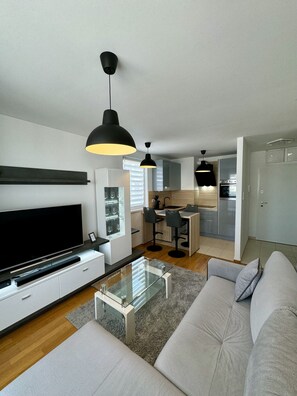 En moderne stue med et fladskærms-tv, et glas sofabord og en grå sofa. Der er et køkken i baggrunden med et spiseområde.