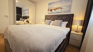 A modern bedroom with a large bed, a headboard with a geometric pattern, bedside lamps, and a painting of a lighthouse on the wall. - Apartment Restart in Zadar (Zadar)