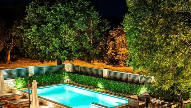 Una piscina rettangolare con lettini e un'area relax, circondata da alberi e da una recinzione.