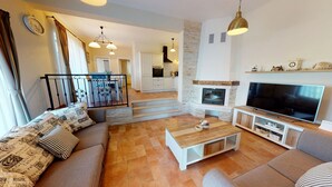 Ein modernes Wohnzimmer mit Sofa, Couchtisch, Fernseher und Kamin. Im Hintergrund ist eine Küche zu sehen und eine Treppe führt zu einer weiteren Ebene.