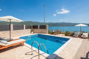 Ein Pool mit freiem Blick auf das Meer und die Berge, umgeben von Liegestühlen und Sonnenschirmen.