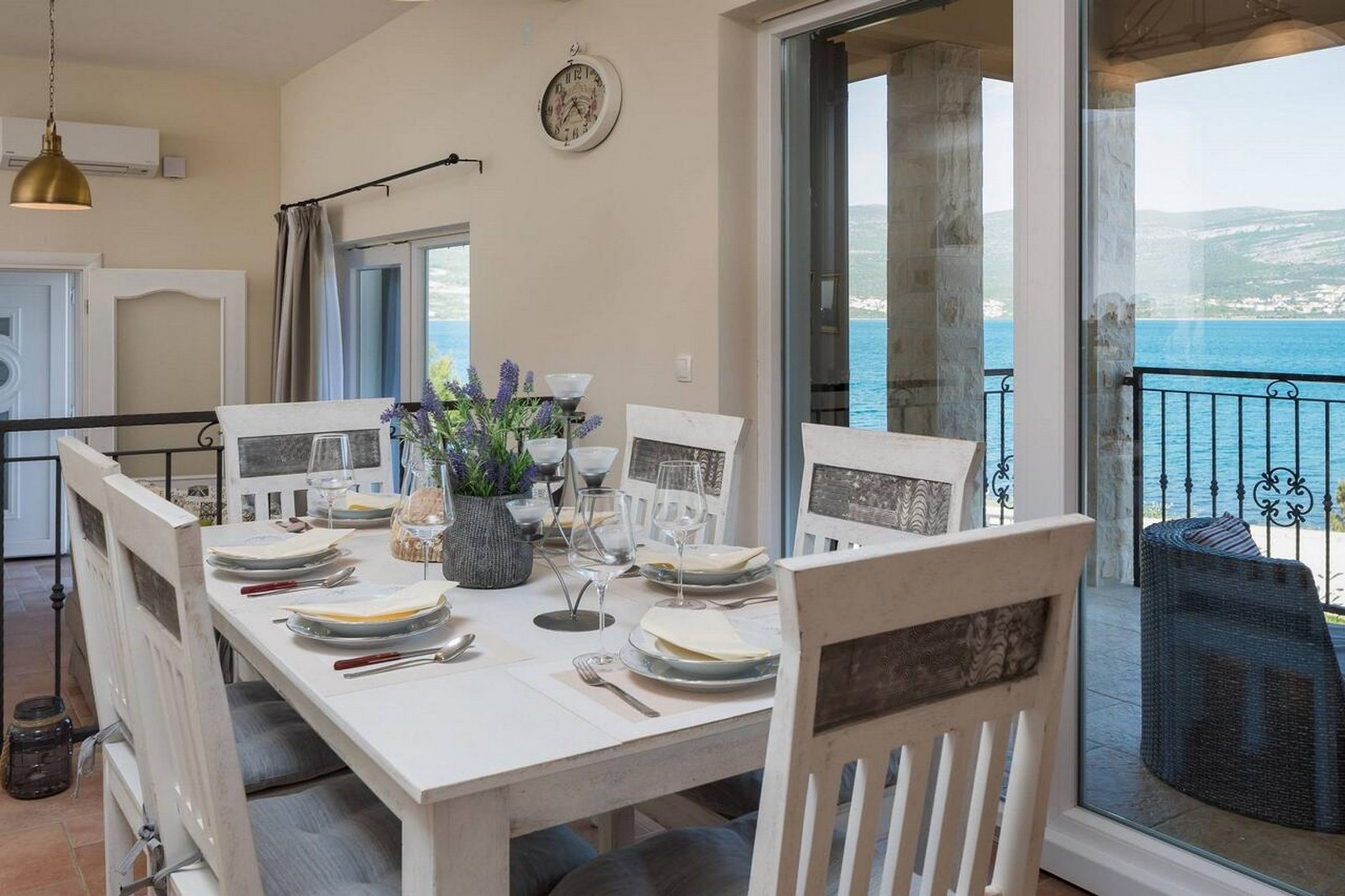 Un coin repas avec une table dressée pour quatre personnes, une horloge au mur et une vue sur la mer par de grandes fenêtres.