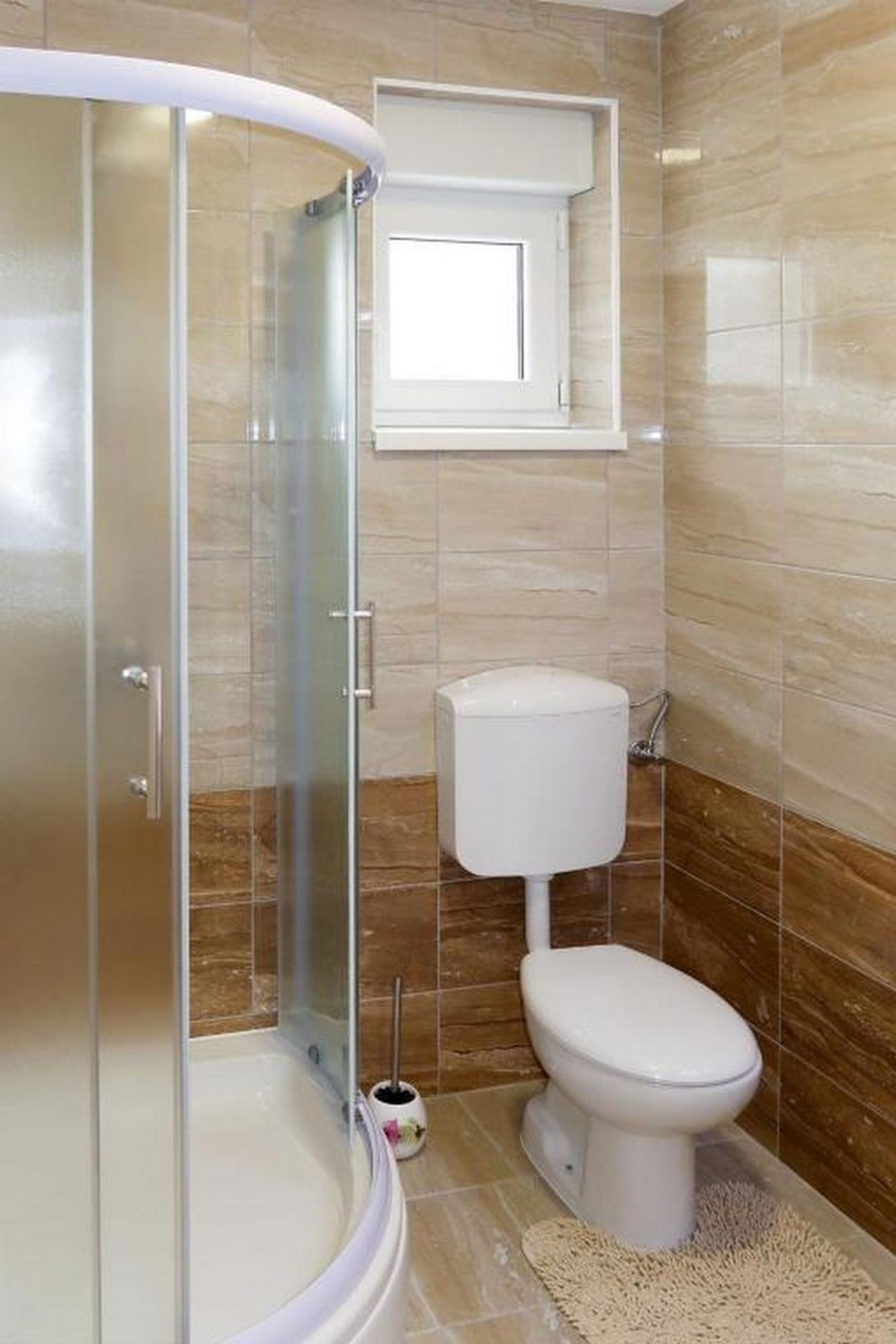 Baño con ducha de vidrio, inodoro y pequeña ventana sobre el inodoro.