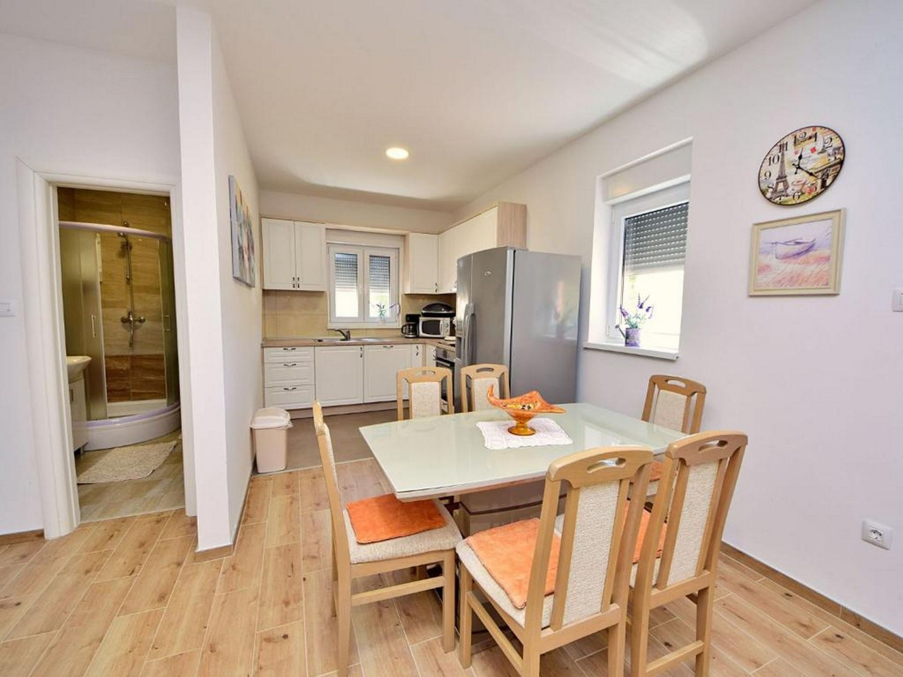 A compact kitchen with white cabinets, a stainless steel refrigerator, and a microwave. There is a dining table with four chairs, and a glass shower enclosure is visible in the background.