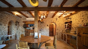 Interior rústic de pedra amb taula i cadires de fusta, parets de pedra i zona de cuina amb armaris de fusta.