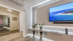 Una moderna hall d'albergo con una grande televisione che mostra una spiaggia, una reception e un divisorio in vetro che conduce alla cucina.