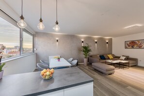 A modern living room with a grey sofa, a dining table with chairs, and a kitchen area with a fruit bowl. - Deluxe Suite Rosa in Zadar (Zadar)