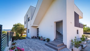 Un bâtiment moderne à deux étages, doté d’un toit plat, d’un escalier menant à l’entrée et d’un balcon agrémenté de plantes en pot.