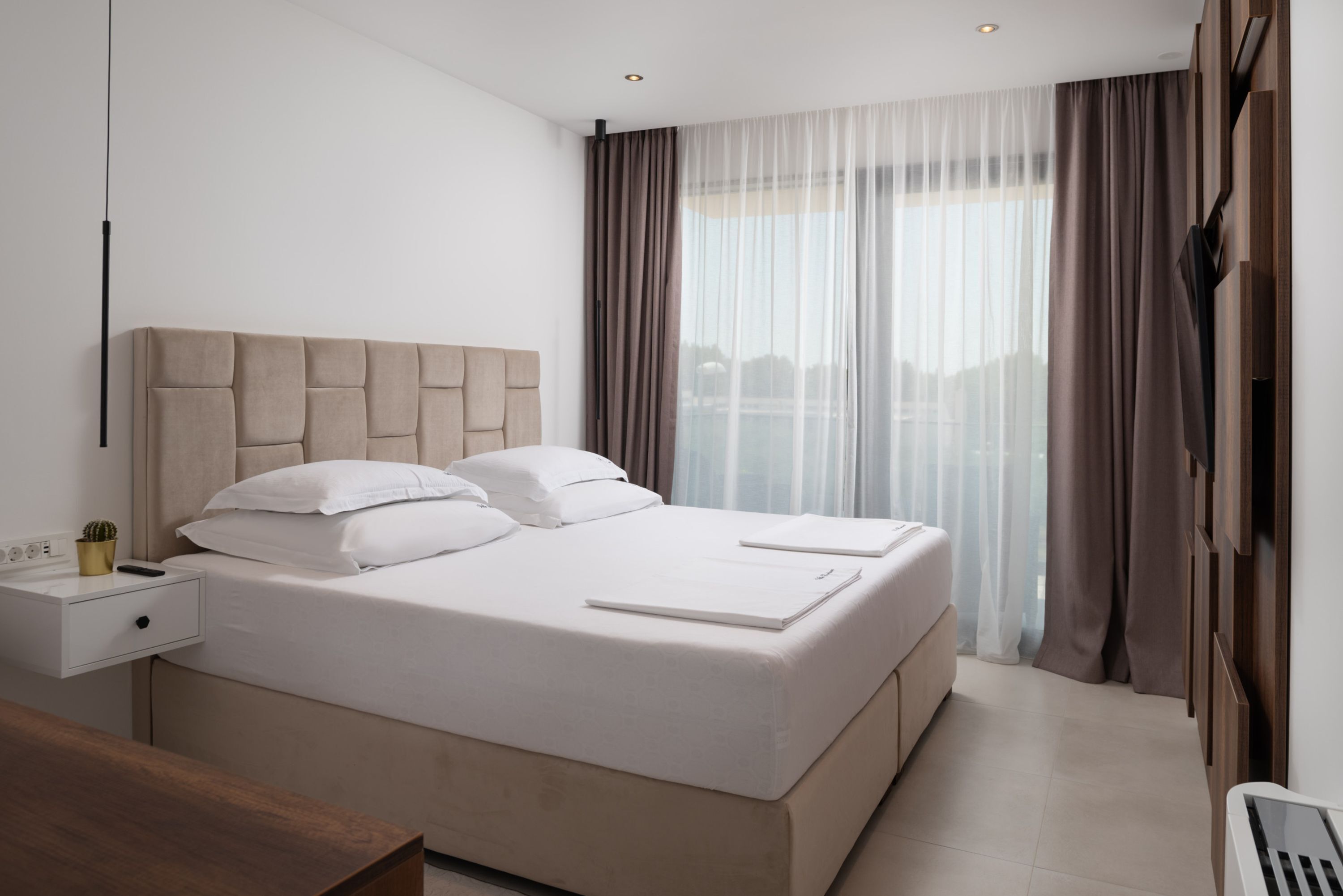 A modern hotel room with a large bed, a bedside table, a nightstand, and a view of the city through sheer curtains.