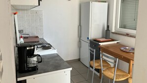 A compact kitchen with a gas stove, a white refrigerator, a small dining table with two wicker chairs, and a window with blinds. - Holiday Home Bella 1 Bella 2 (Povljana)