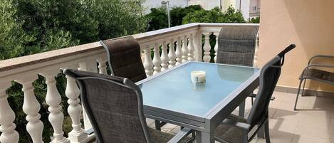 A balcony with a glass table and four chairs, a white balustrade, and a view of houses in the background.