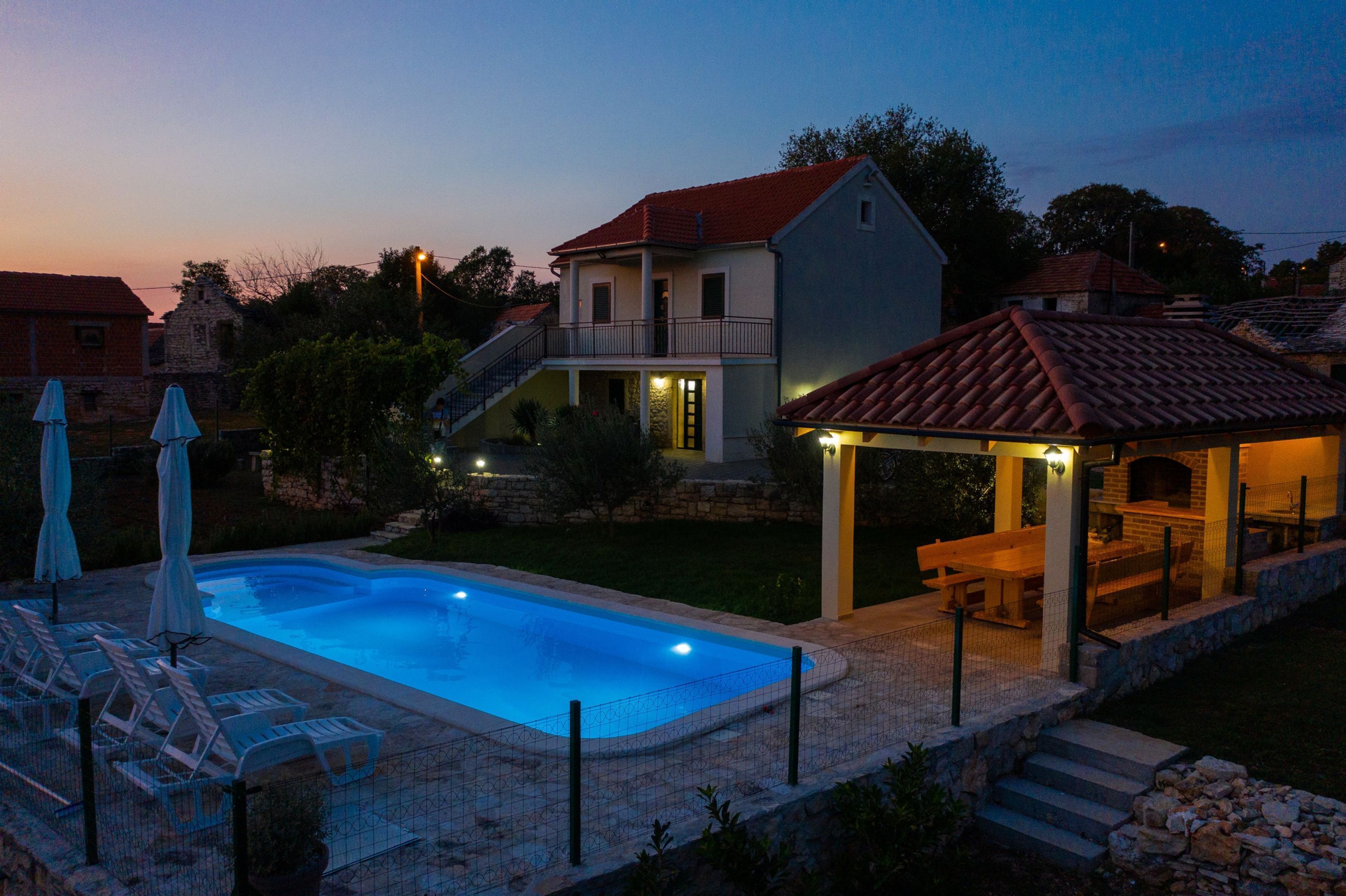 A swimming pool with lounge chairs, a covered patio with a table and chairs, and a stone staircase leading to a house with a red roof.