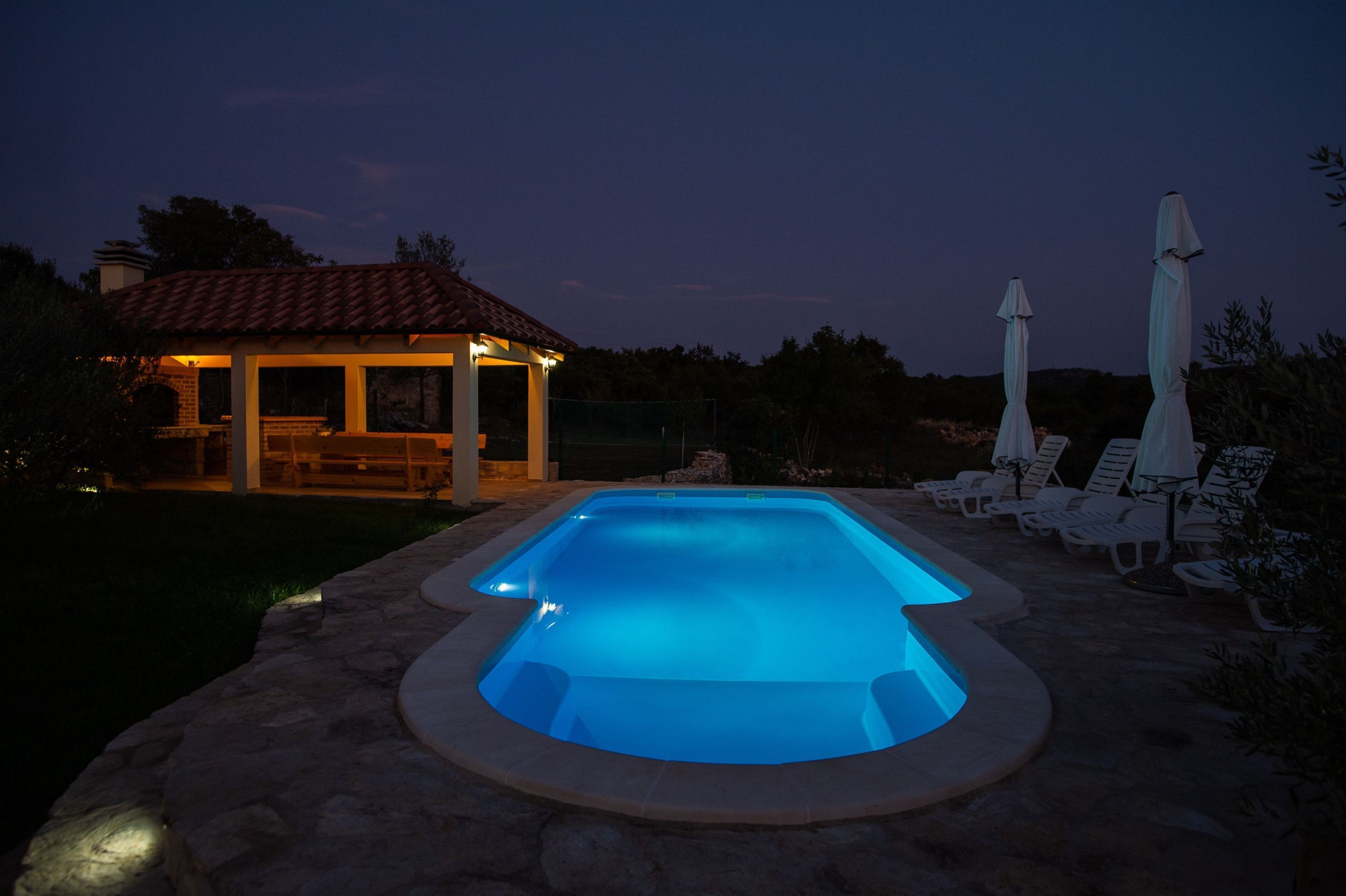 A swimming pool with a stone deck, surrounded by lounge chairs and umbrellas, and a covered seating area with a tiled roof.