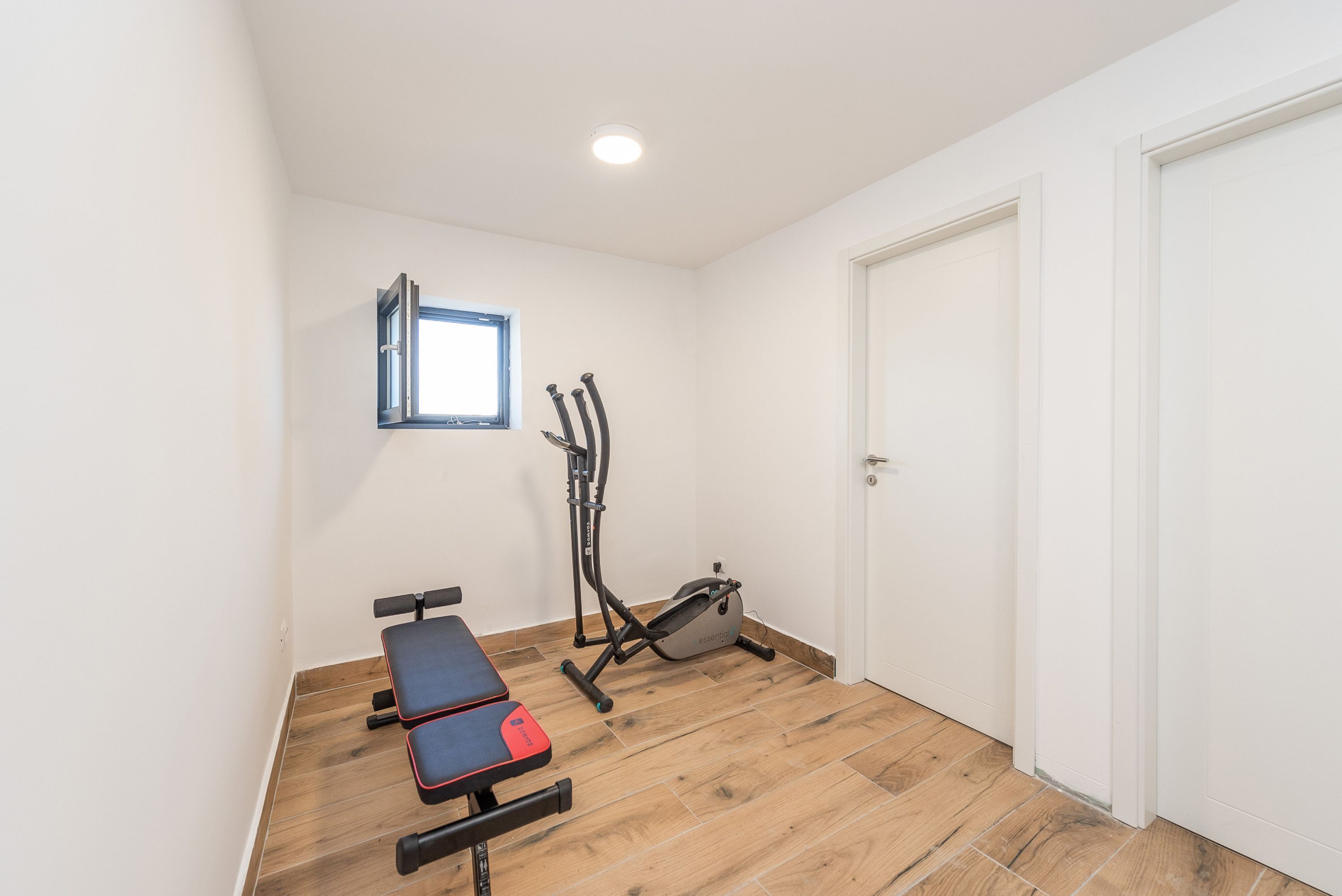 Ein kleiner, gut beleuchteter Raum mit Holzboden, einer Bank, einem Ergometer und einer Tür.