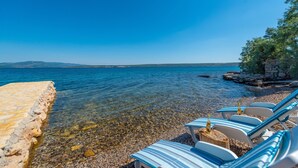 A beachside view with clear waters, a stone wall, and striped lounge chairs with bottles. - Beachfront Villa Promenada (Posedarje)