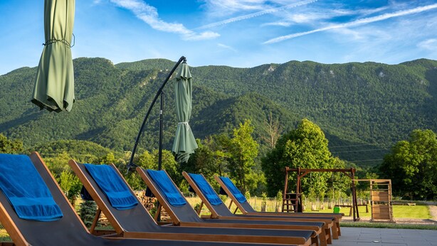 Una hilera de sillones de madera con toallas azules, un conjunto de columpios de madera y vistas a montañas verdes bajo un cielo despejado.