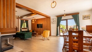 Un amplio salón con chimenea, vigas de madera, un sofá verde, un sillón mecedora amarillo y una zona de comedor con mobiliario de madera.