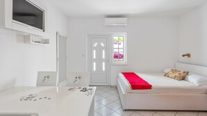 A minimalist room with a bed, a TV mounted on the wall, and a window with a view of pink flowers. - Superior Studio Tania 16 (Sveti Filip i Jakov)