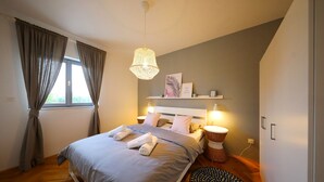 Ein Schlafzimmer mit Bett, Nachttischlampen, einem Nachttisch, einem Fenster mit Vorhängen und einem an der Wand befestigten Regal.