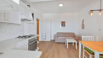 A modern kitchen with white cabinetry, a gas stove, and a wooden floor. There is a dining area with a table and chairs, and a sofa in the background.