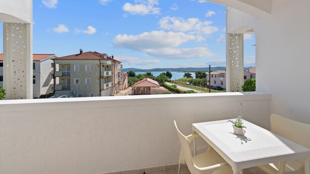 A balcony with a table and chairs, overlooking a residential area with buildings and a body of water. - Superior Studio Tania 3 (Sveti Filip i Jakov)