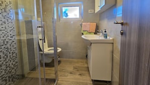 A bathroom with a glass shower enclosure, a toilet, a sink with a soap dispenser, and a window above the sink. - Studio Apartment Nia (Nin)